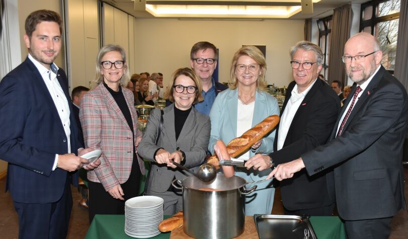 Kartoffelsuppe-Essen Handwerkspräsident, Kreishandwerkermeister, Geschäftsführer und drei Damen als Gäste stehen um einen Topf mit Würstchen und Brot